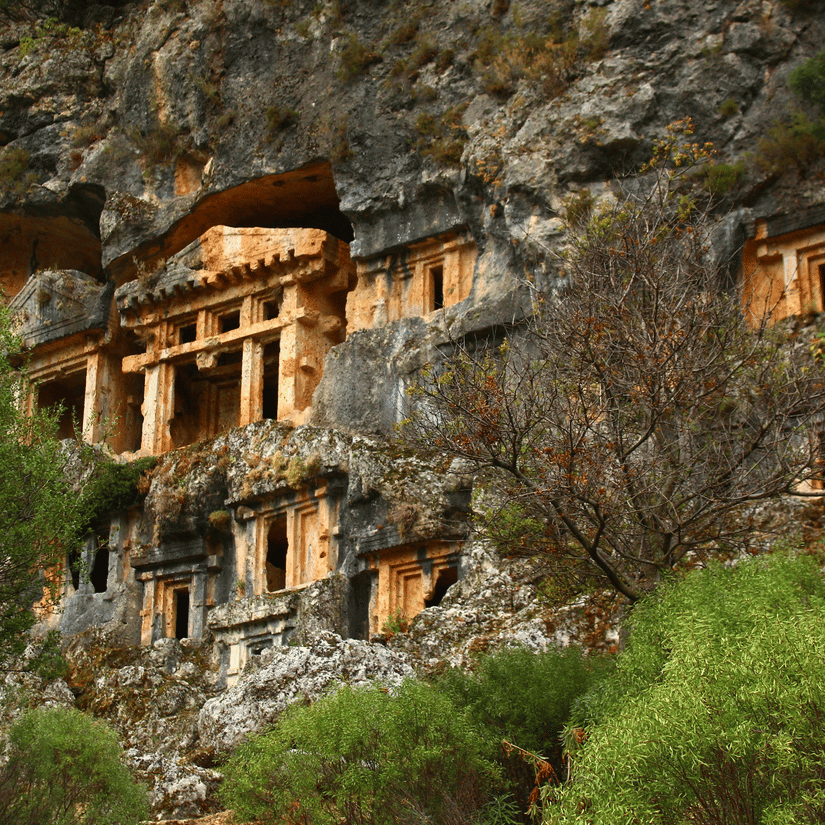 A view from afar of a caves cut from the side of a cliff with windows, doors, and pillars in view.