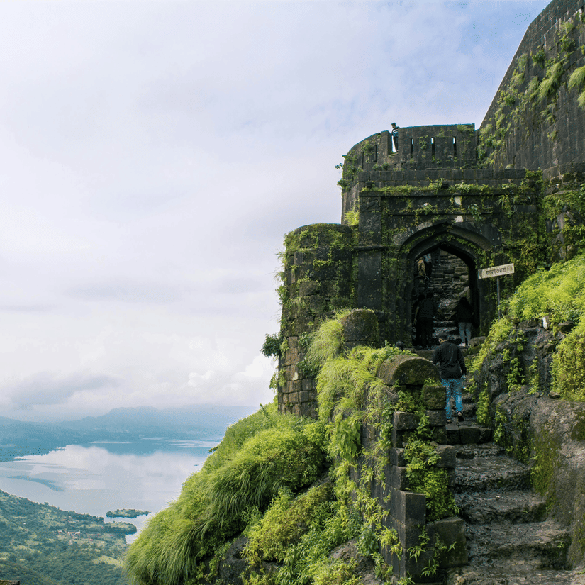 A view from afar of the steps covered by moss and grass on the way to Lohagadh Fort in Lonavala with a lake and clouds seen in the distance.