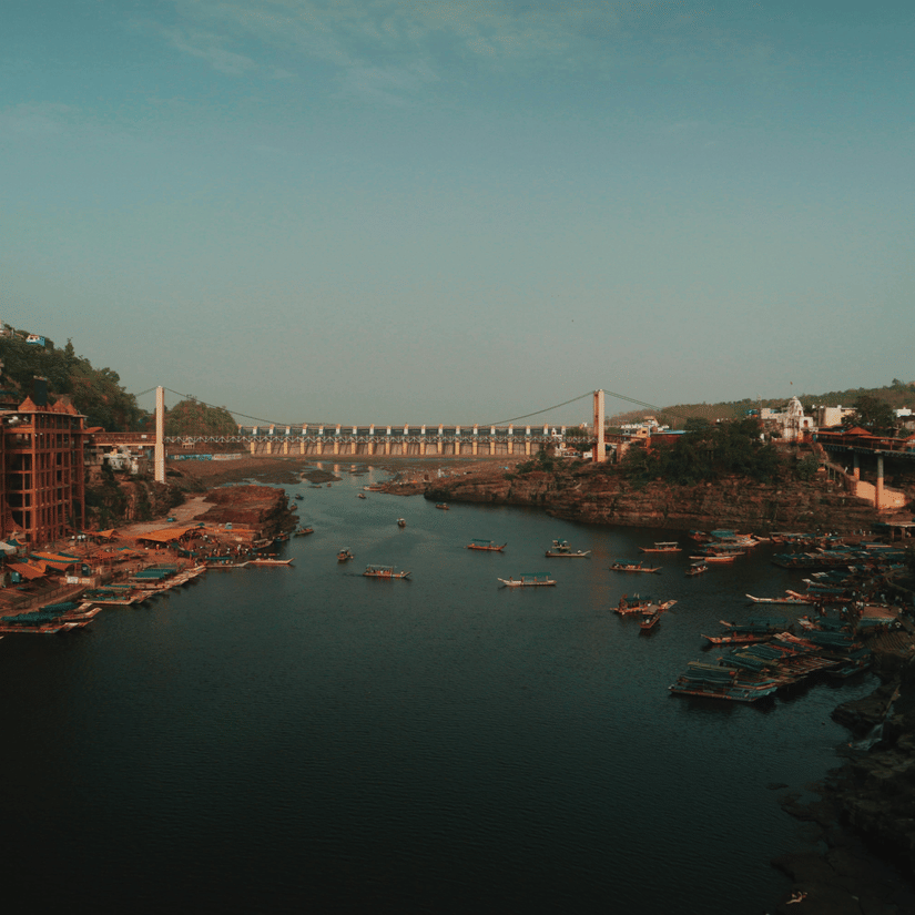 A far-out view of Omkareshwar Temple on the banks of Narmada River with a bridge in the centre and boats on the river. Omakareshwar is one of the main stops on Narmada Parikrama by foot.