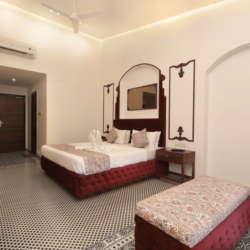 Elegant hotel bedroom with double bed, patterned bedding, bedside lamps, decorative mirror with arches, cushioned bench, tiled floor, and warm ambient lighting.