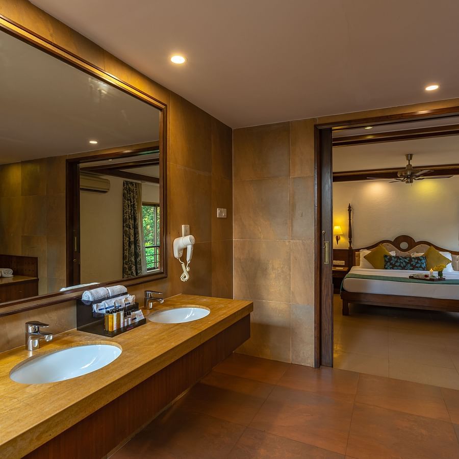 An image of a washroom featuring a large square mirror, a sink, towels, and shower amenities, with an open door revealing a room that includes a bed.