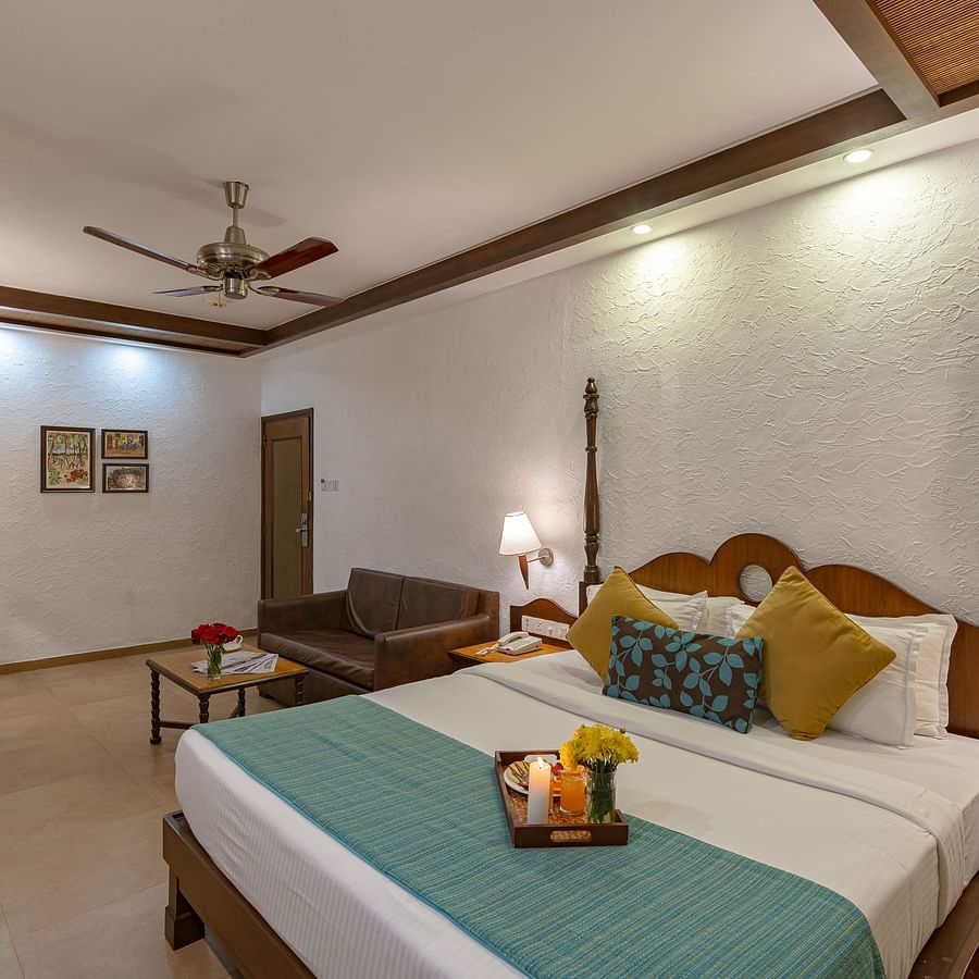 An image of a room with a sofa, table lamps, a vanity, wall paintings, and a neatly made bed featuring a tray with a candle, juice, flowers, and snacks.