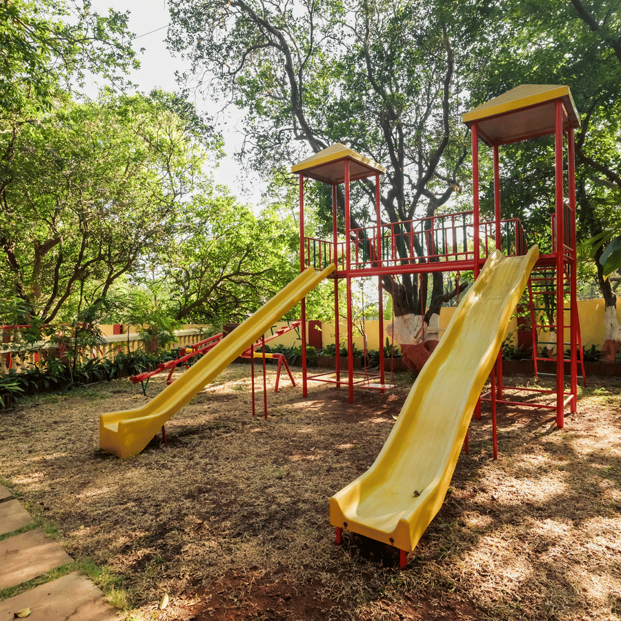 Outdoor children's play area with slides set under large trees at Adamo The Village.