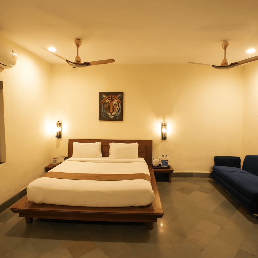 A well-lit resort bedroom with a double bed centered, a side window, and a long blue sofa couch.