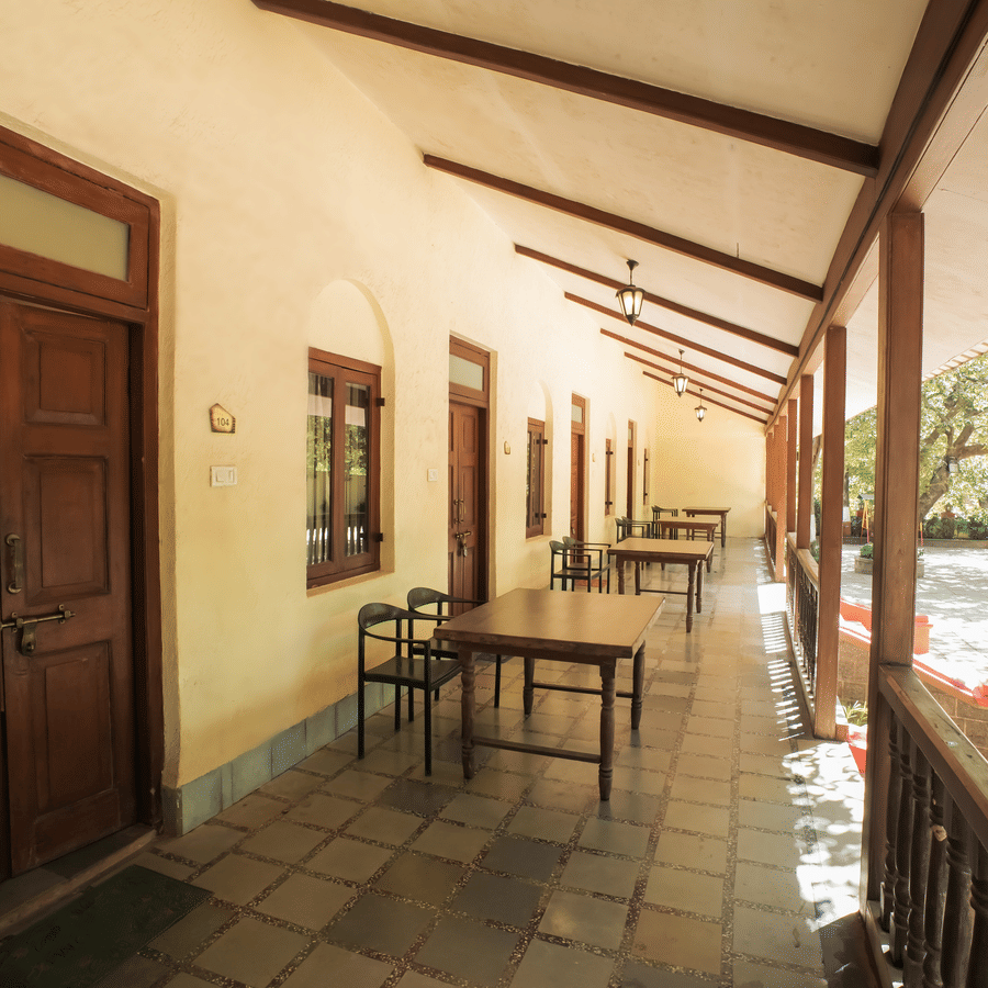 A long outdoor veranda corridor with wooden doors, pillars, and small seating tables overlooking a garden area.