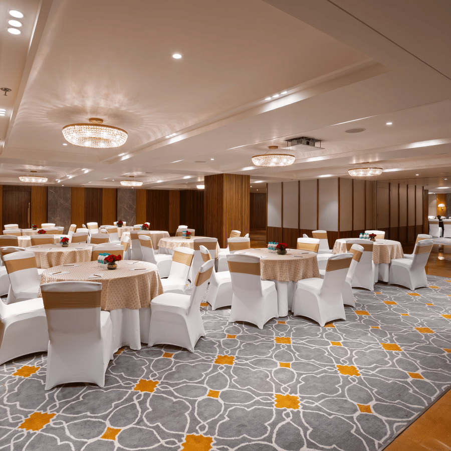 Spacious banquet hall at Aloha on the Ganges Resort with round tables, white chairs, patterned carpet, wood-paneled walls, chandeliers, and podium for events.