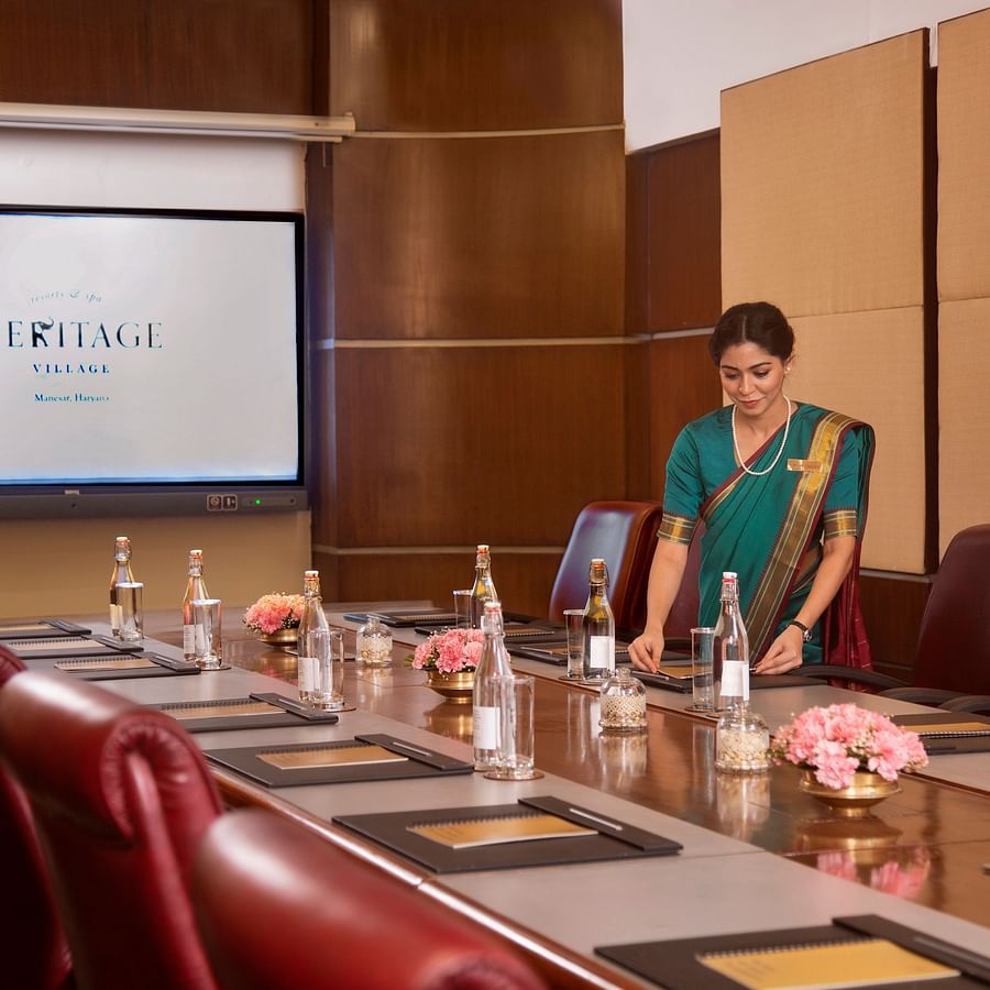A modern boardroom setup featuring leather chairs, a wooden conference table with notepads, water bottles, floral decor, and a large presentation screen