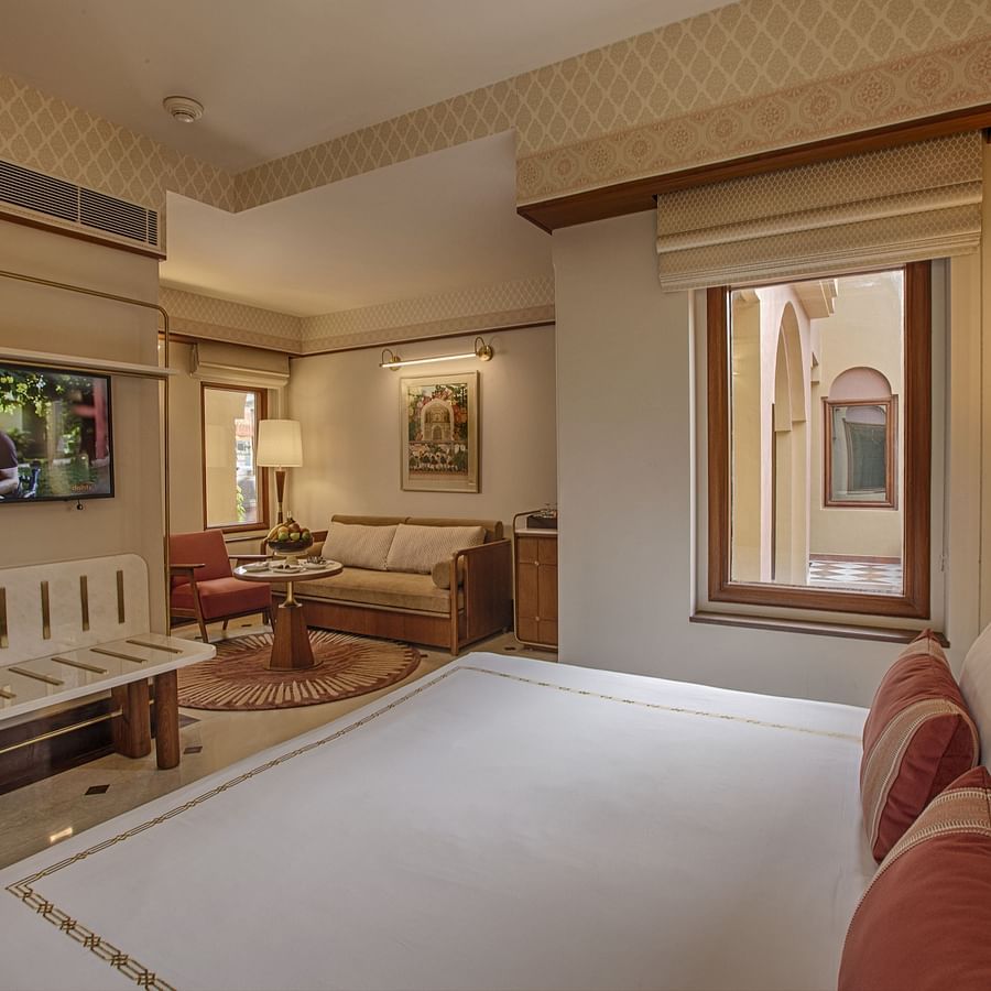 A hotel bedroom with a neat bed, wall-mounted TV, small window, and a seating area with a sofa, chair, and coffee table - Heritage Village Resorts & Spa, Manesar