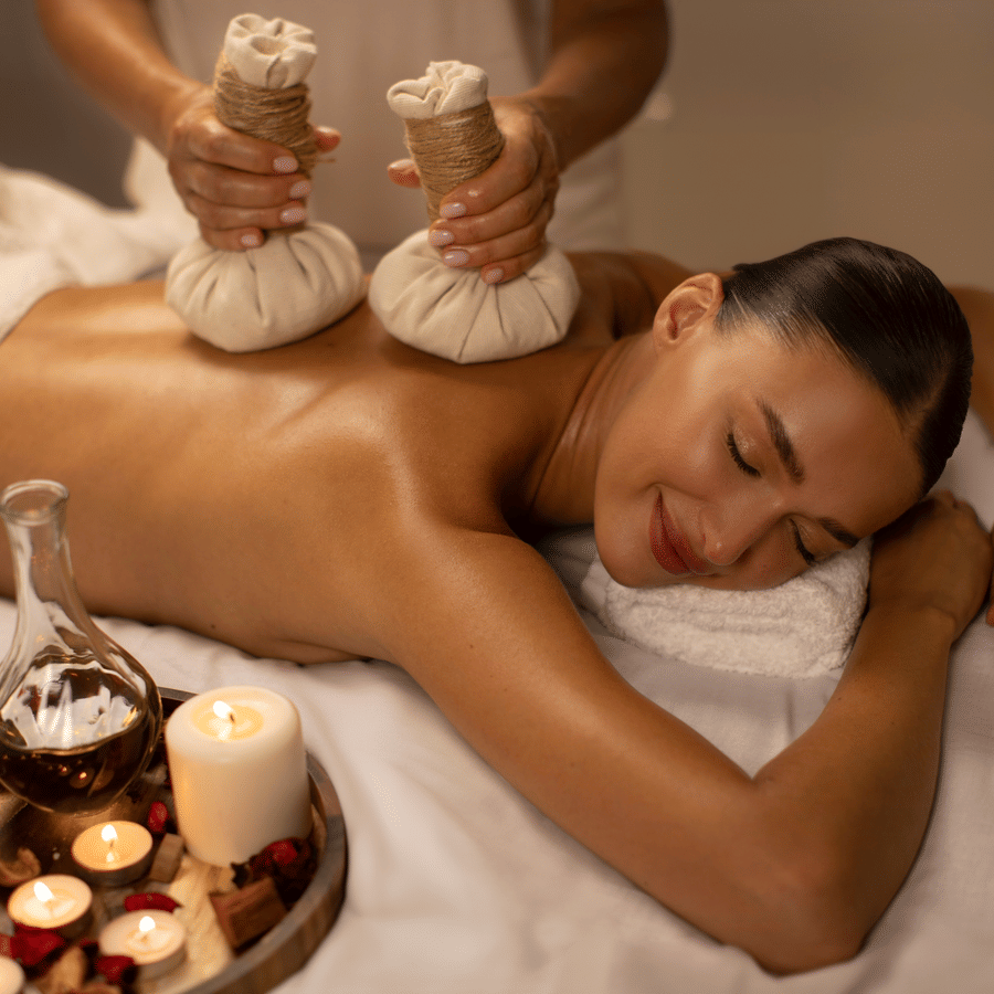 Woman receiving a herbal compress back massage surrounded by spa candles and oils.