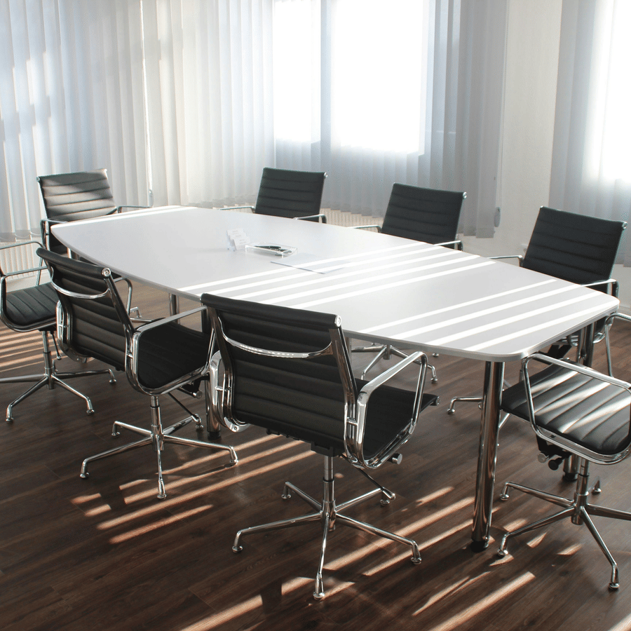 Modern conference room with white rectangular table, eight black office chairs, large windows with vertical blinds, and dark wooden flooring, designed for meetings and discussions.