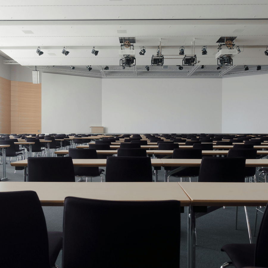 Spacious lecture hall with rows of light-colored tables and black chairs, white ceiling with mounted lights and projectors, podium at front, and modern design for presentations.