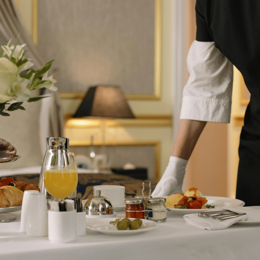 Hotel staff serving in-room breakfast with croissants, juice, flowers, and a plated hot dish on a white table cloth.
