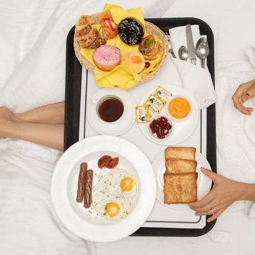 In-room breakfast tray with juice, fruits, and pastries placed beside a person on the bed at Heritage Village Resorts & Spa, Goa.