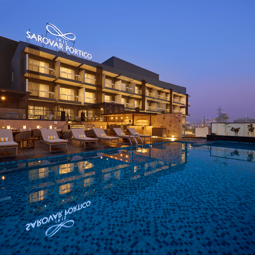 A modern hotel building is illuminated at dusk, featuring a rooftop swimming pool reflecting the hotel's signage and nearby lounge chairs. - Iris Sarovar Portico, Sector 85, Gurugram