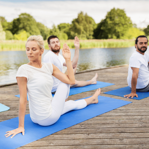 Group of people practicing seated spinal twist yoga poses on mats by a lakeside wooden deck, surrounded by greenery and calm water