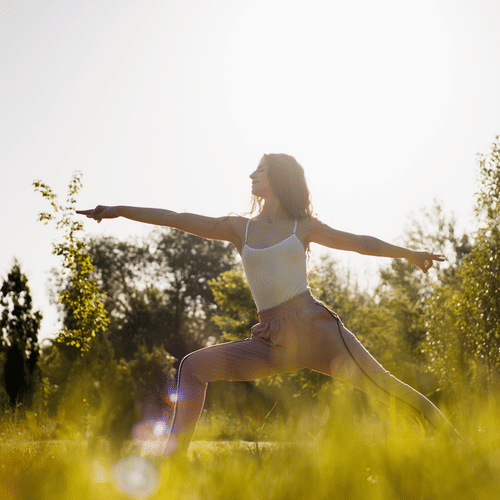 Woman practicing yoga outdoors in a grassy field, holding Warrior II pose with arms extended, surrounded by sunlight and trees