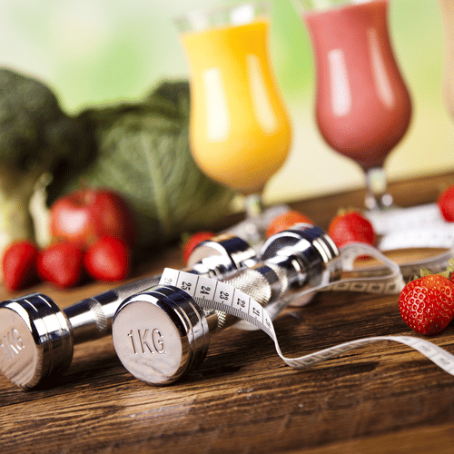 A set of 1 kilo dumbbells on a wooden surface with a set of vegetables and glasses of juices in the background