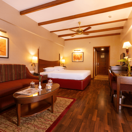 A bedroom at The Carlton in Kodaikanal featuring a double size bed, a work desk and chair, TV screen, a sofa and centre table.