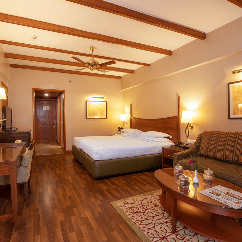 A bedroom at The Carlton featuring a bed, work desk and chair, lamps, seating area with sofa and a flat TV screen, one of the cosiest Kodaikanal rooms.