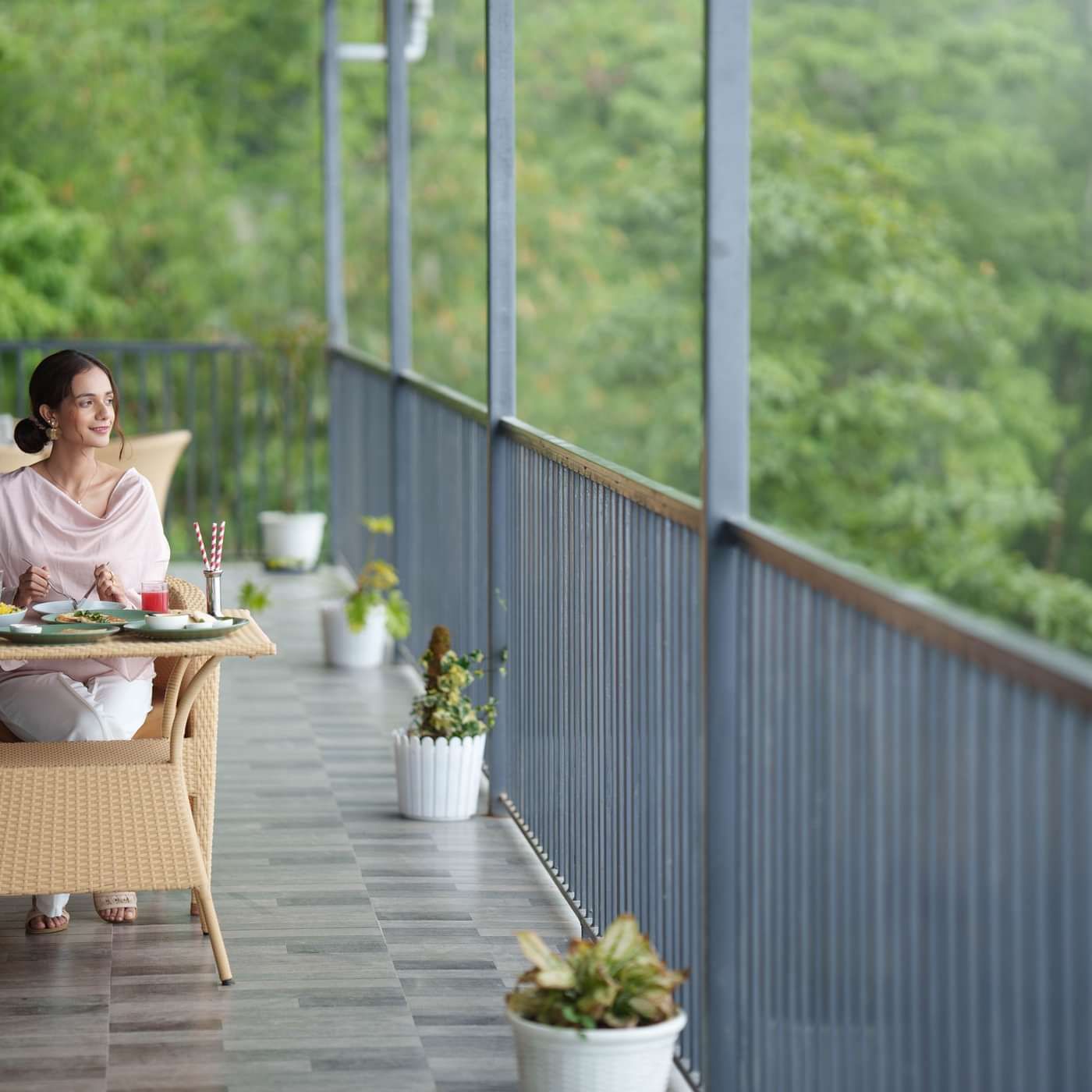 A woman in a light pink outfit enjoys a meal at a wicker table on a balcony, surrounded by potted plants and overlooking lush green trees.