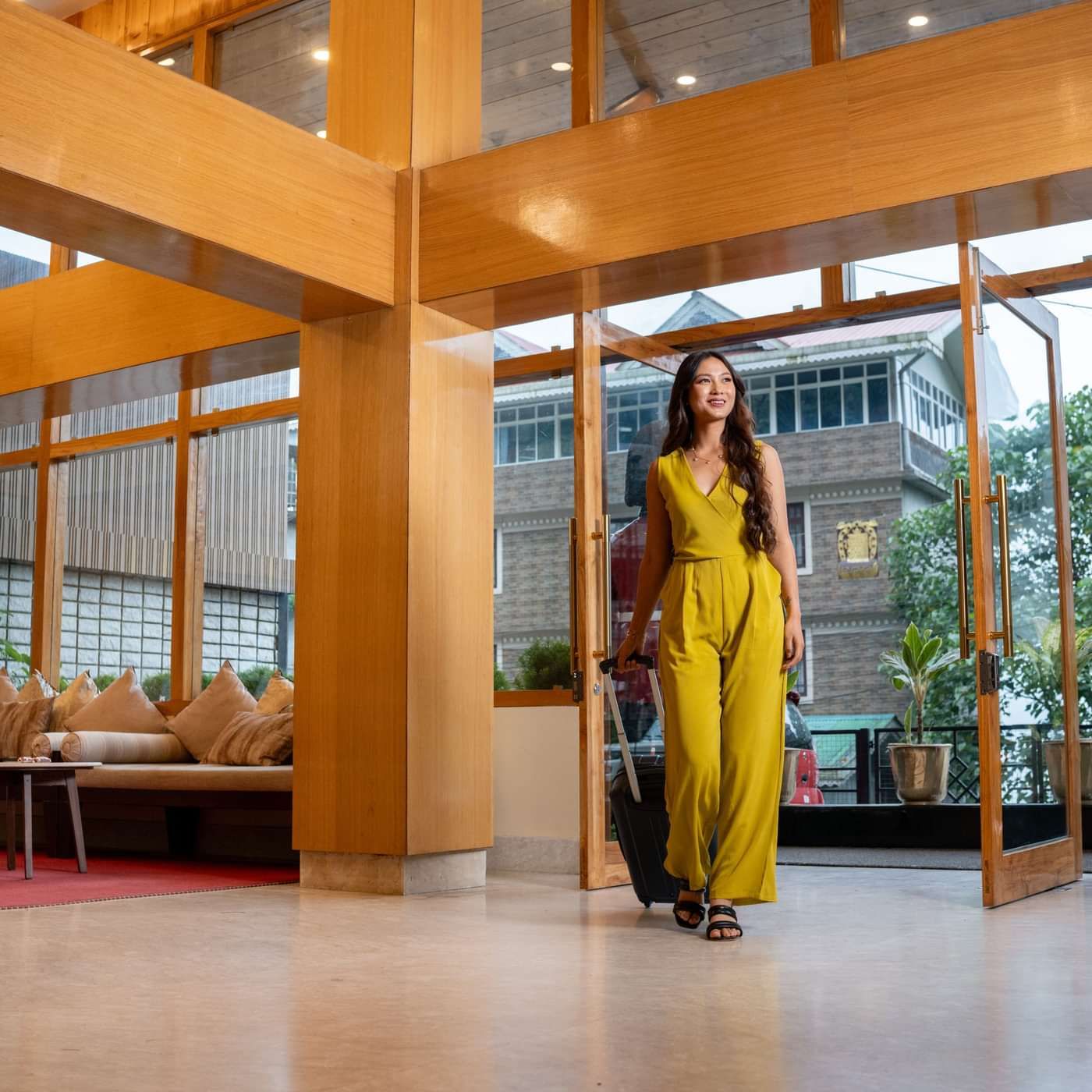 A woman in a yellow jumpsuit walks into a hotel lobby with a suitcase, passing through glass doors. The lobby has wooden frames, seating, and outside greenery.