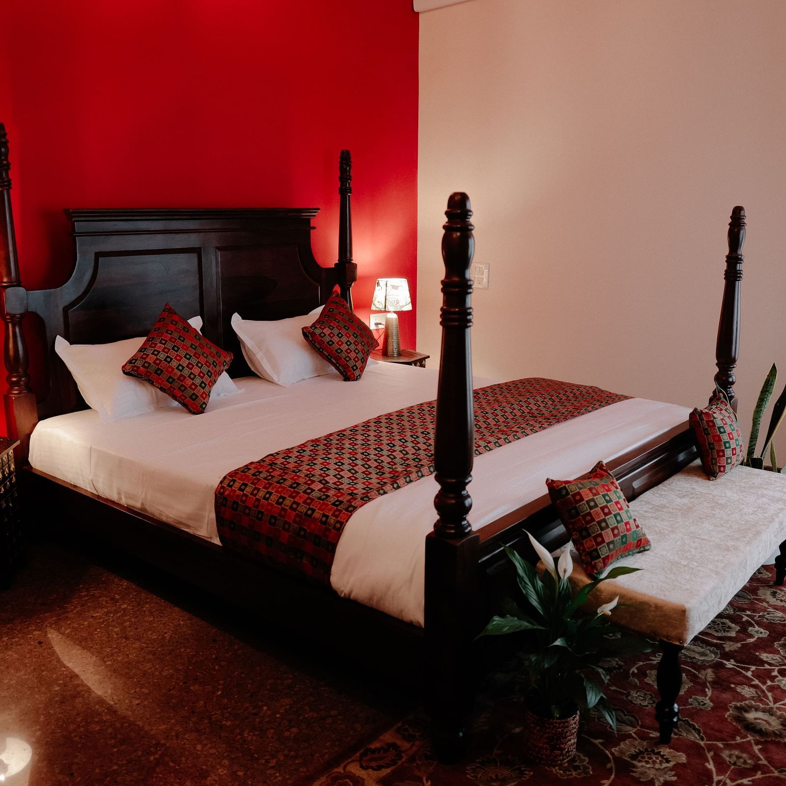 A dark wood four-poster bed with white linens and red patterned throw pillows in a room with a bold red accent wall and a lighter adjacent wall | Adhyaya Heritage Reverbed