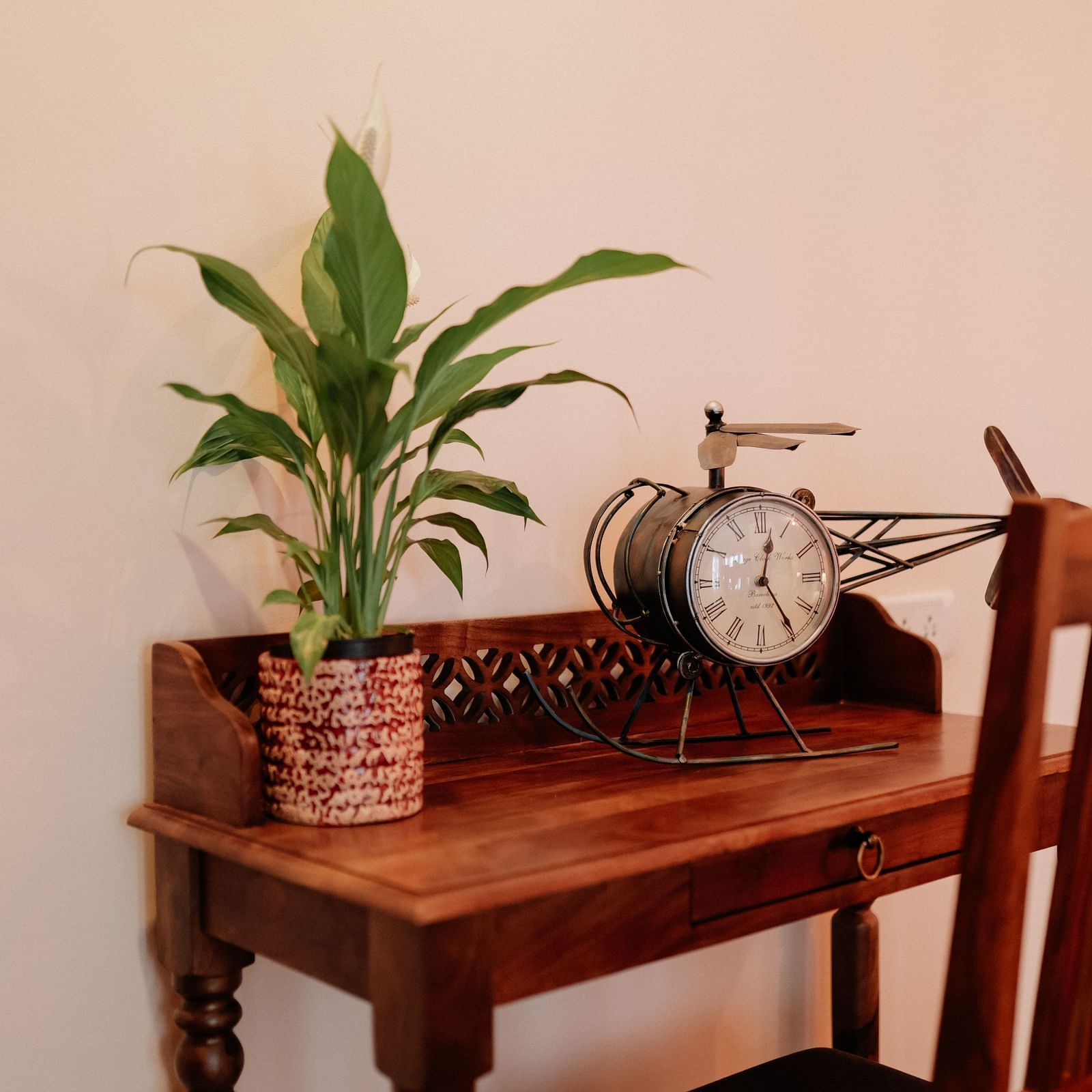 A wooden desk with a potted plant and a vintage clock on top, with a wooden chair beside it, against a plain wall | Adhyaya Heritage Reverbed
