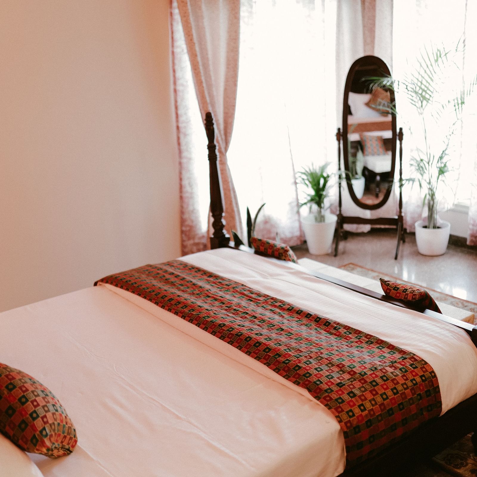 A bedroom with a wooden four-poster bed, white linens, patterned throw pillows and a blanket, and a leaning mirror with plants in the background  | Adhyaya Heritage Reverbed