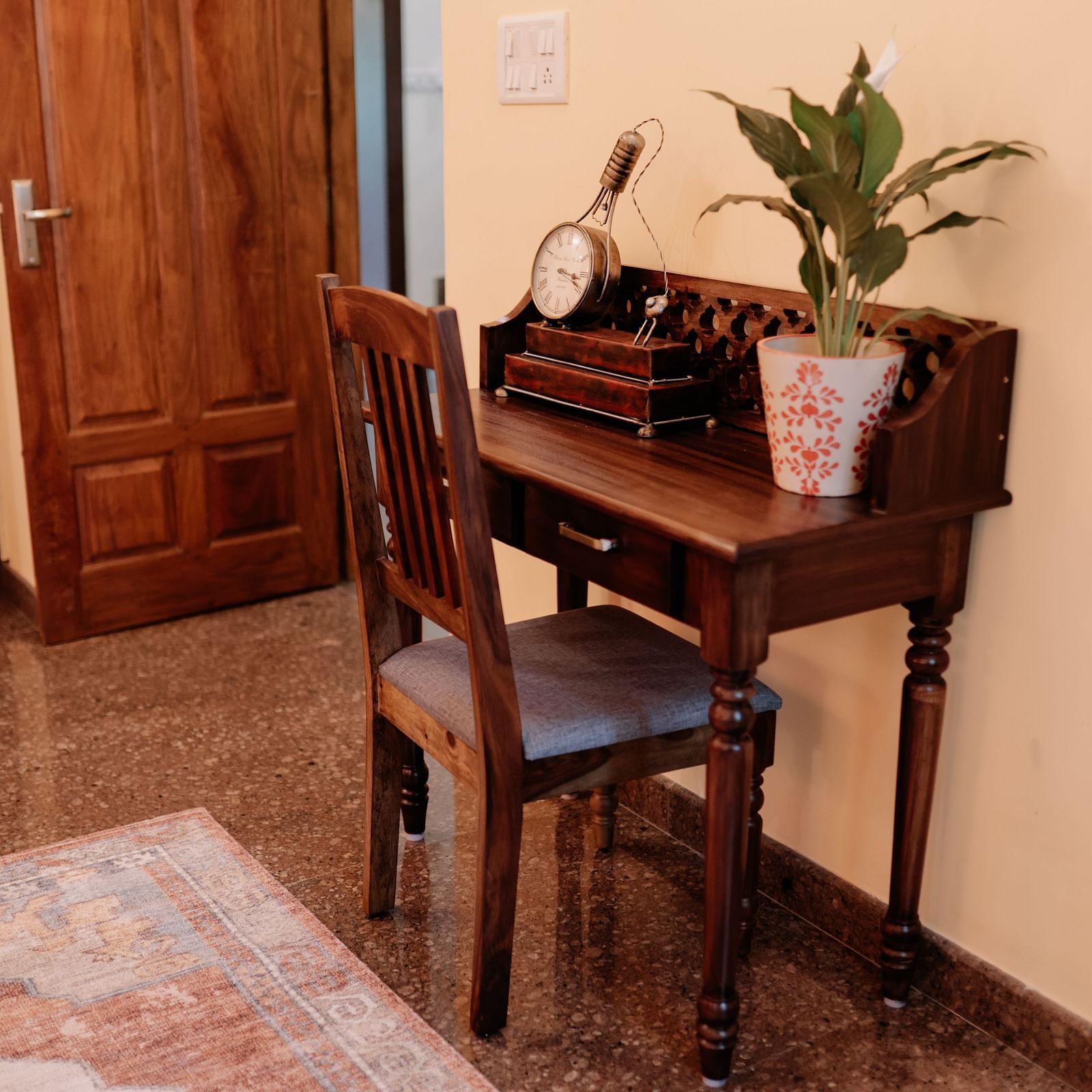 A wooden desk with a plant, a small clock, and a chair in a hallway with a wooden door | Adhyaya Heritage Reverbed