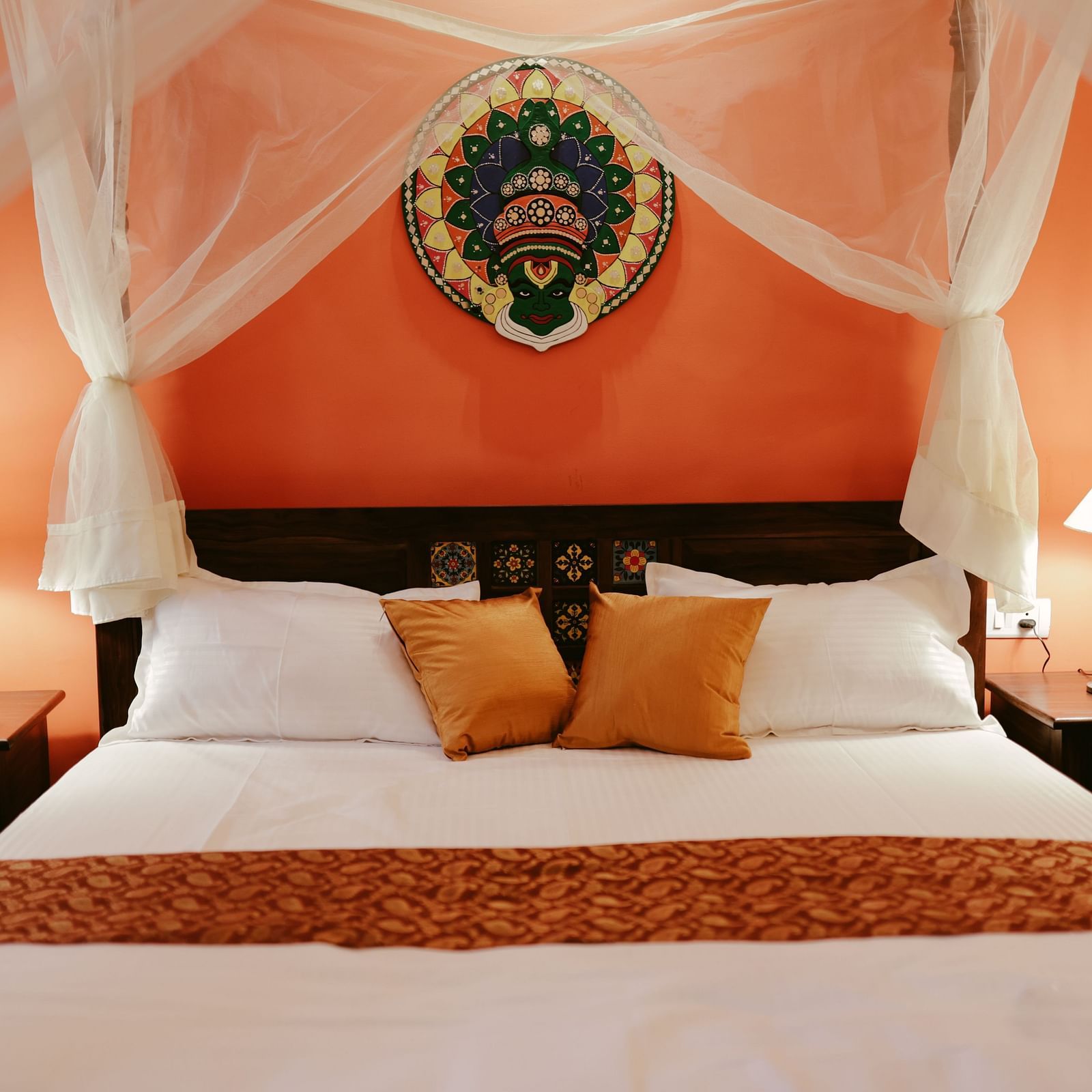 A bed with white linens and orange pillows, flanked by two side tables with lamps, against an orange wall with a decorative plate | Adhyaya Heritage Reverbed