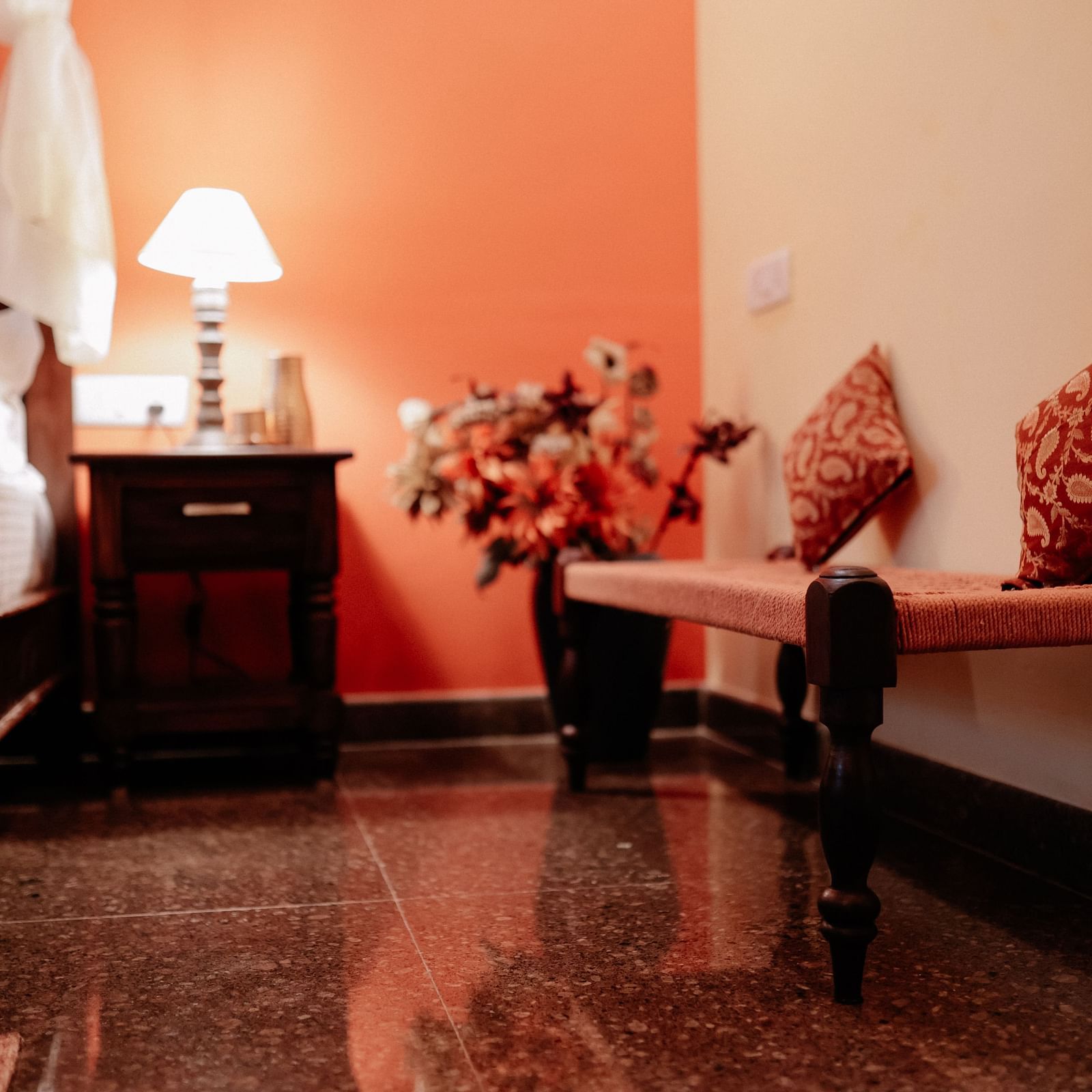 A bedroom corner with a wooden bench with cushions, a side table with a lamp, and a glimpse of a bed with white drapery  | Adhyaya Heritage Reverbed