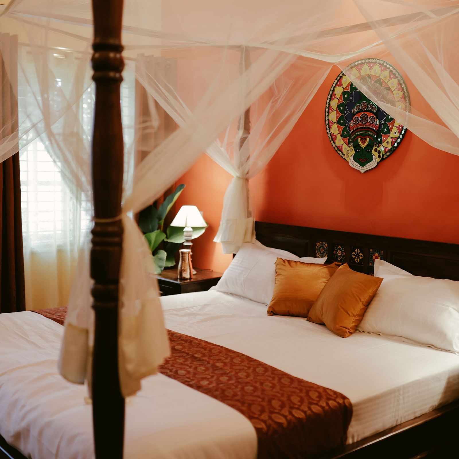A four-poster bed with white drapery and a decorative headboard in a room with an orange accent wall | Adhyaya Heritage Reverbed