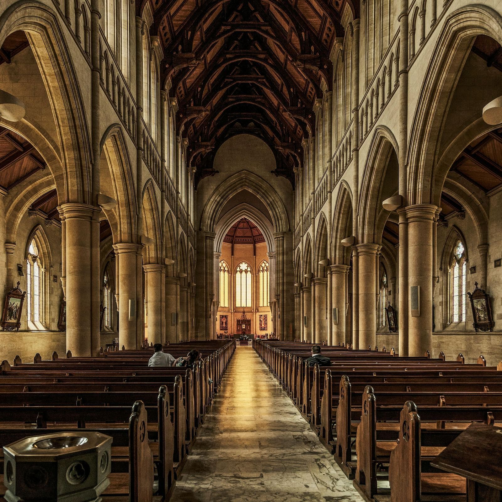 A grand cathedral interior with high vaulted ceilings, stained glass windows, and rows of wooden pews leading to a brightly lit altar.