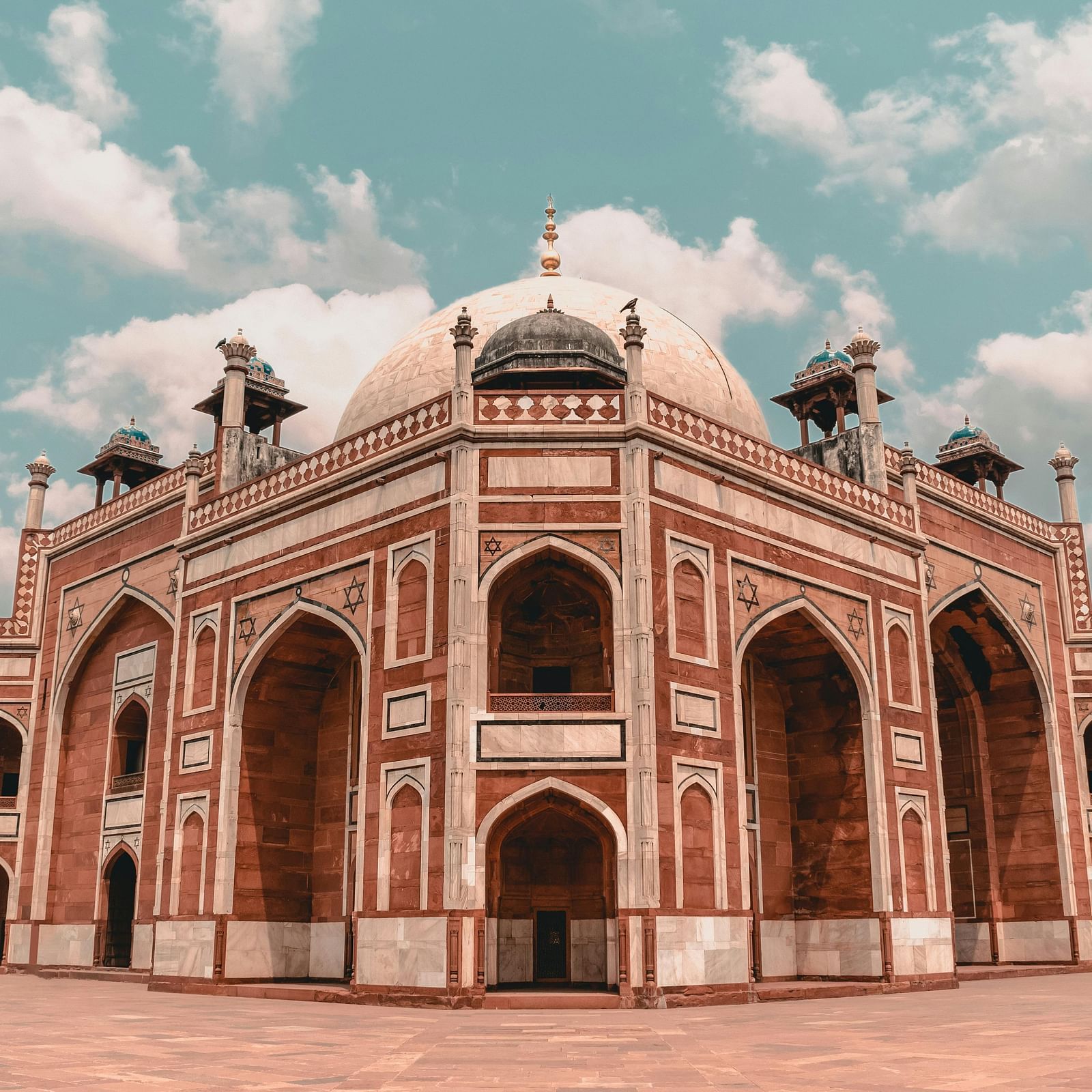 Humayun's Tomb, a grand Mughal-style building with red sandstone and white marble, features arched entrances and domes under a partly cloudy sky.