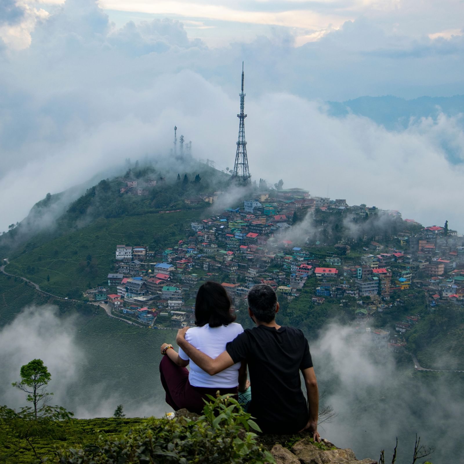 Romantic couple sitting on a hilltop overlooking Darjeeling town covered in clouds and mist, with panoramic Himalayan mountain views — best tourist attraction in West Bengal.
