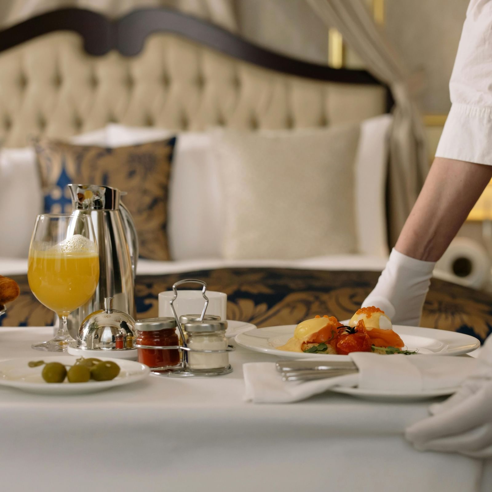 A pair of hands setting up a breakfast tray with orange juice and food on a table in front of the bed.