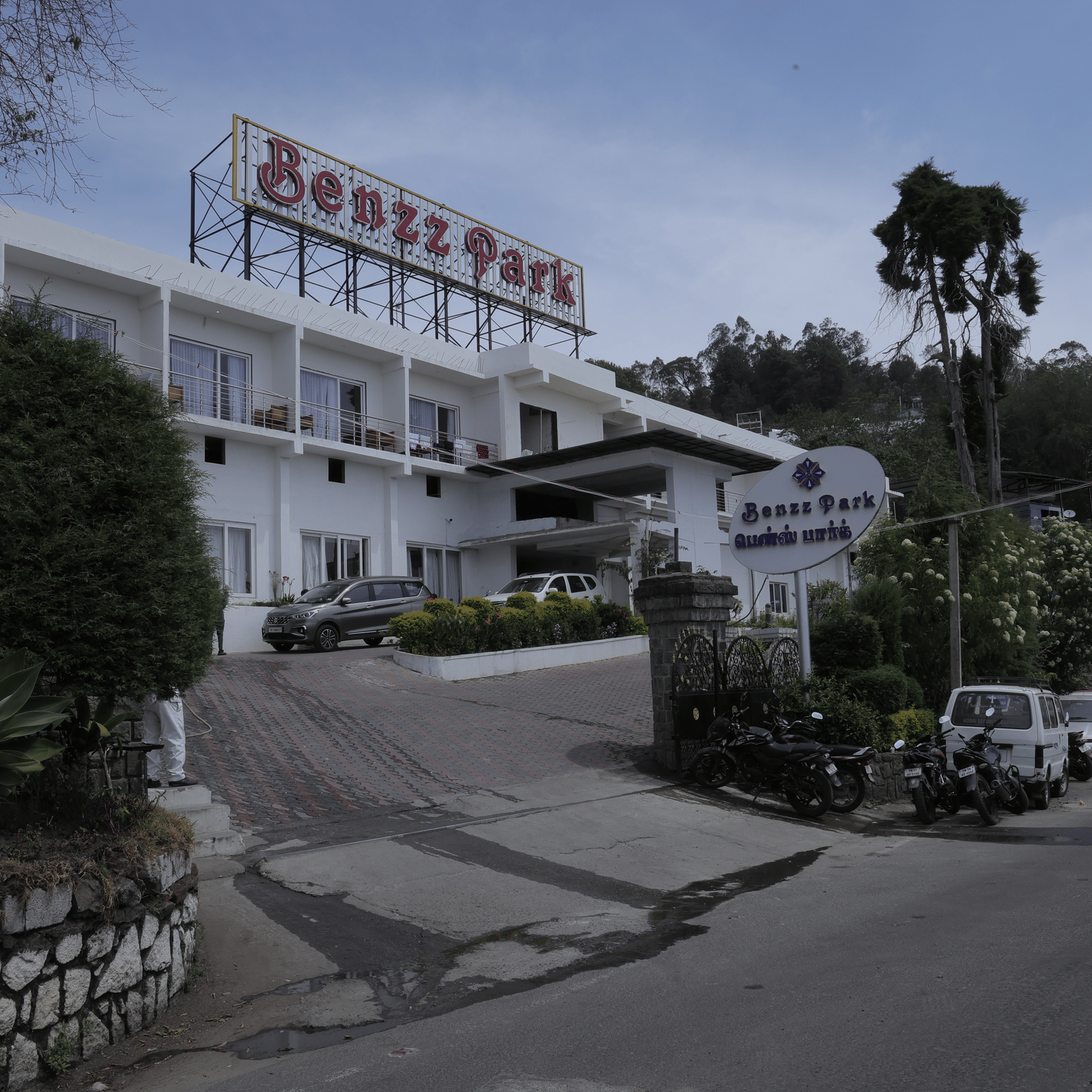 The Benzz Park hotel building exterior on a hillside road, surrounded by trees and vehicles.