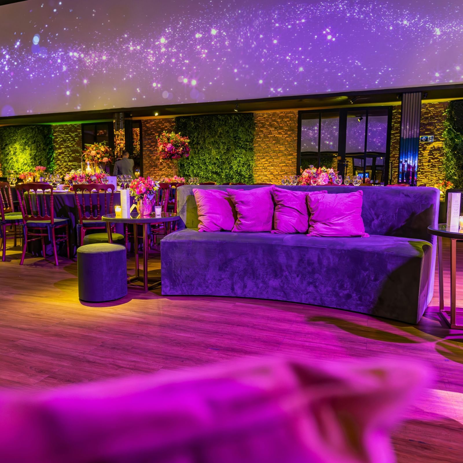 A lounge area for a party, featuring purple couches, vibrant purple and pink lighting, and a large screen displaying an image.