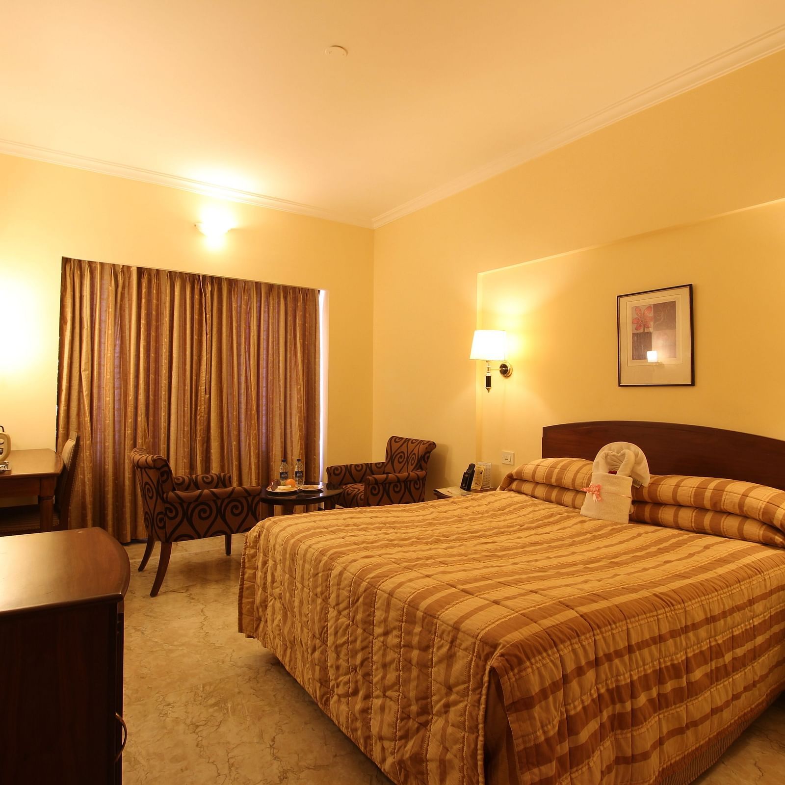 A room with a large bedding with wall lamps on either side of the bed, coffee table and chairs, on a bright lighted setting.