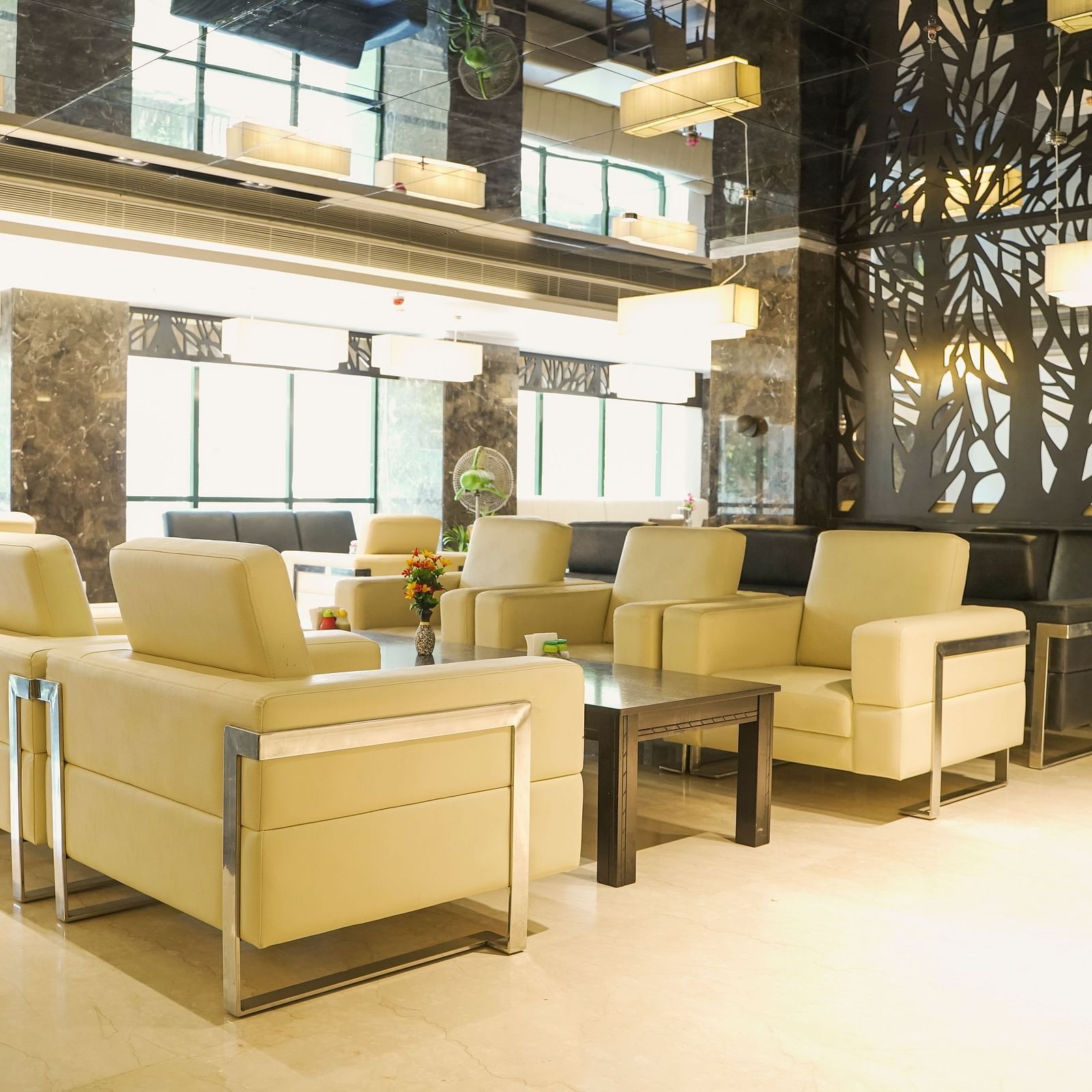 A broad view of a luxurious lounge at BNGV Grandeur Hotel and Banquets, featuring contemporary light-coloured seating, dark decorative wall panels, and natural light from large windows.