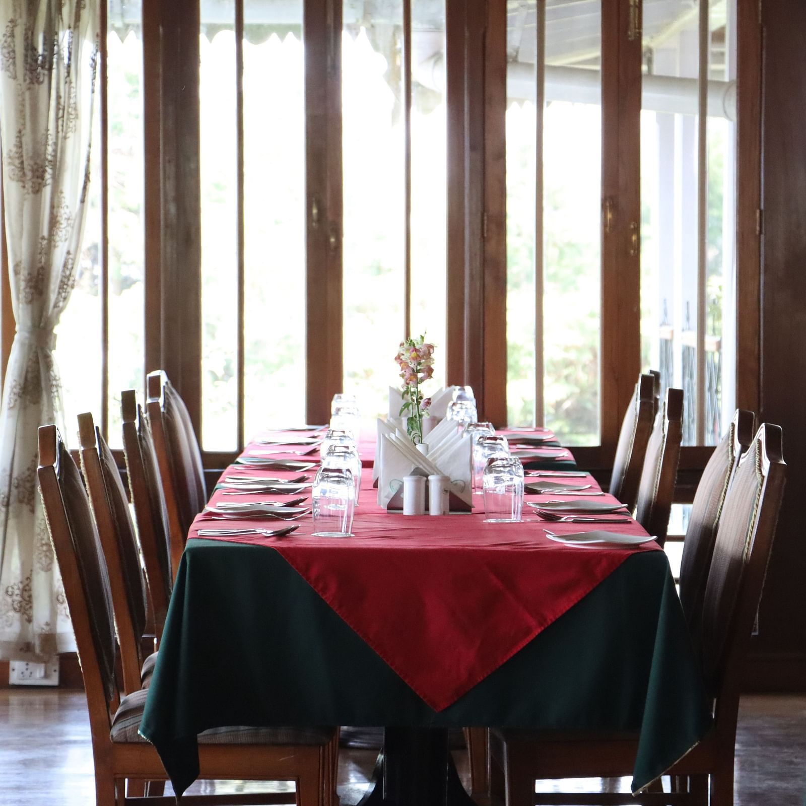 The eight seating table in the dining room of Cedar Inn, Darjeeling placed beside a glass and wood framed wall, arranged with cutlery and other dining amenities.