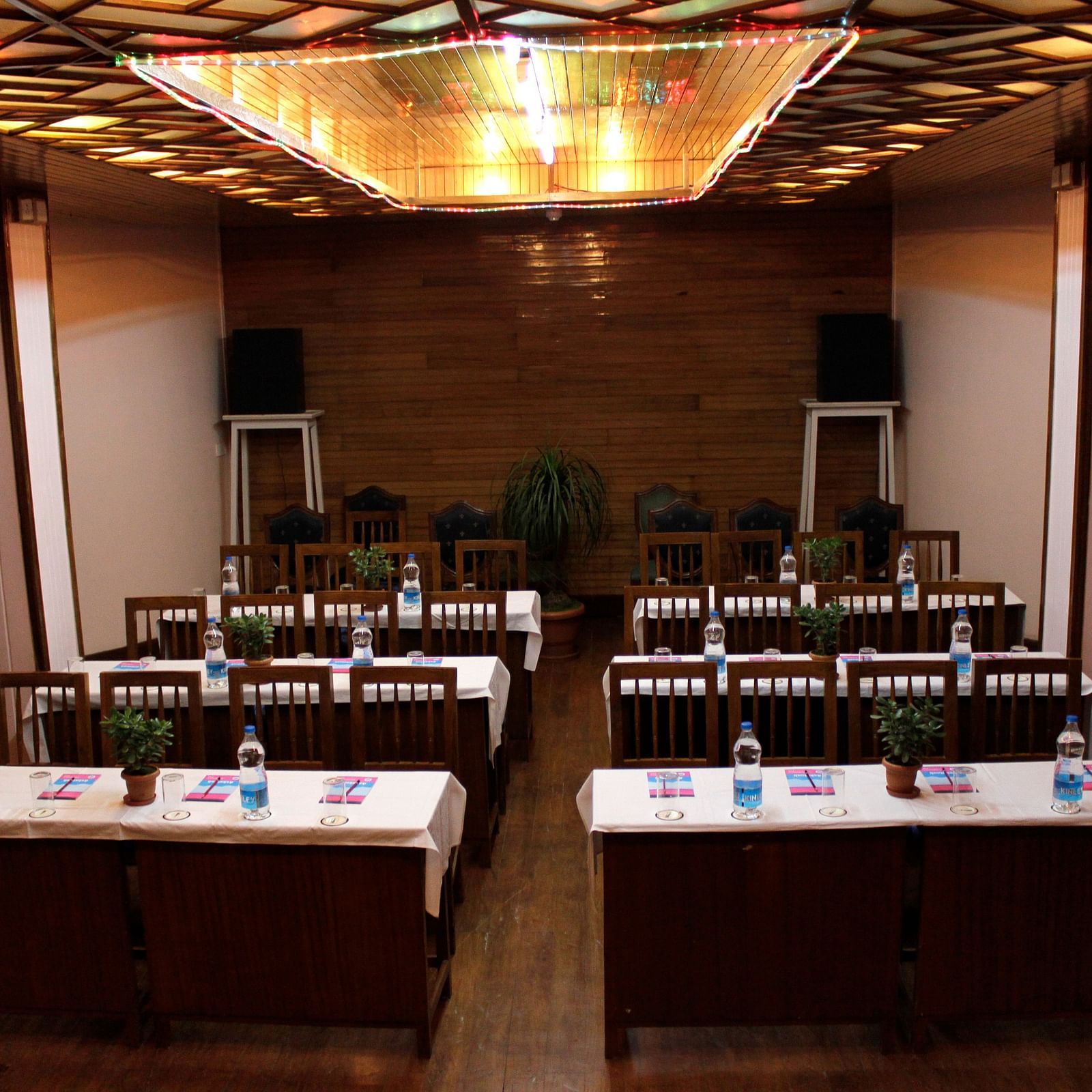 Classroom seating arrangement in the Banquet hall of Cedar Inn, Darjeeling featuring six tables with four chairs each, overhead lighting and speakers.