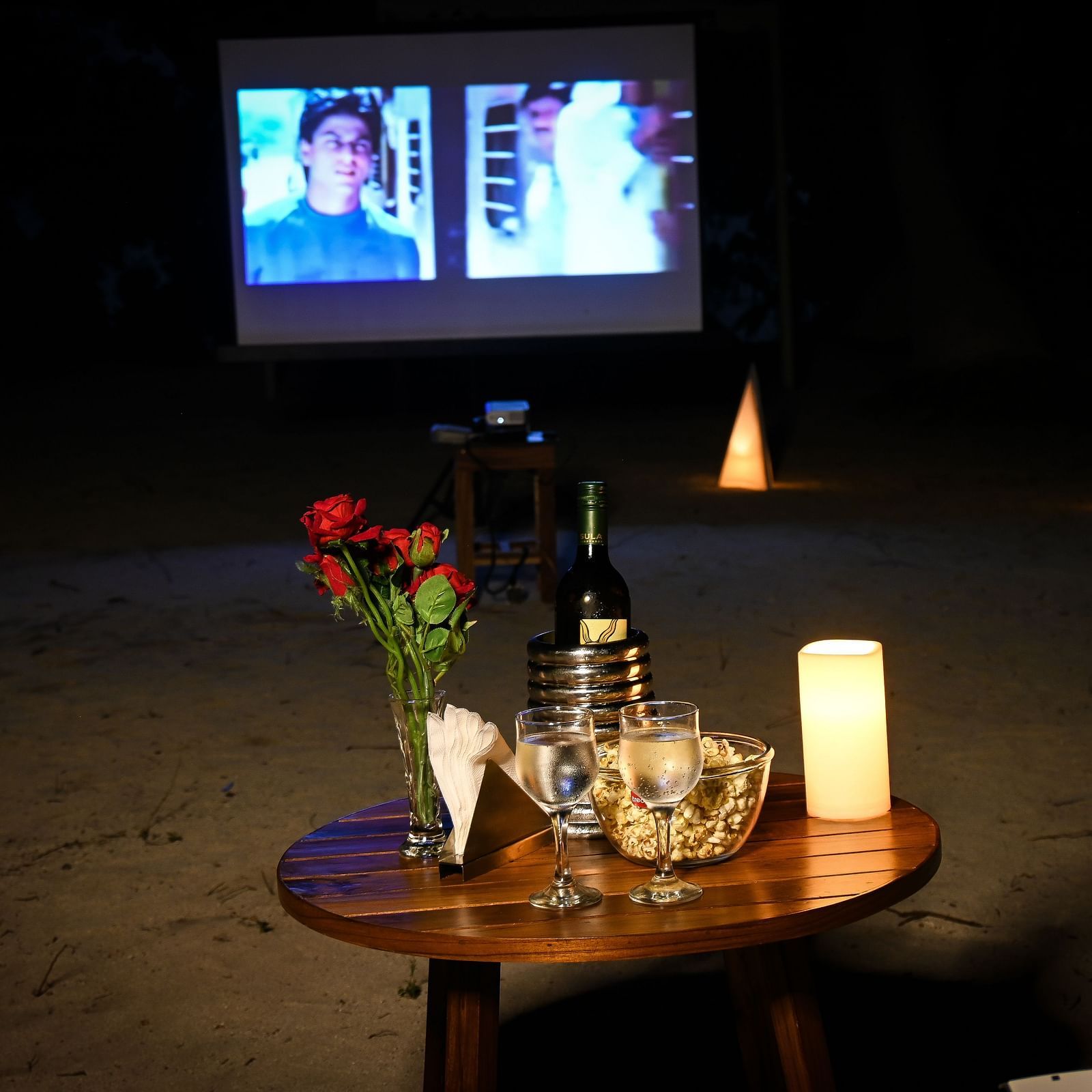 A small table with glasses wine rose on top and a movie playing in the white screen in the background - Coral Reef Resort & Spa, Havelock