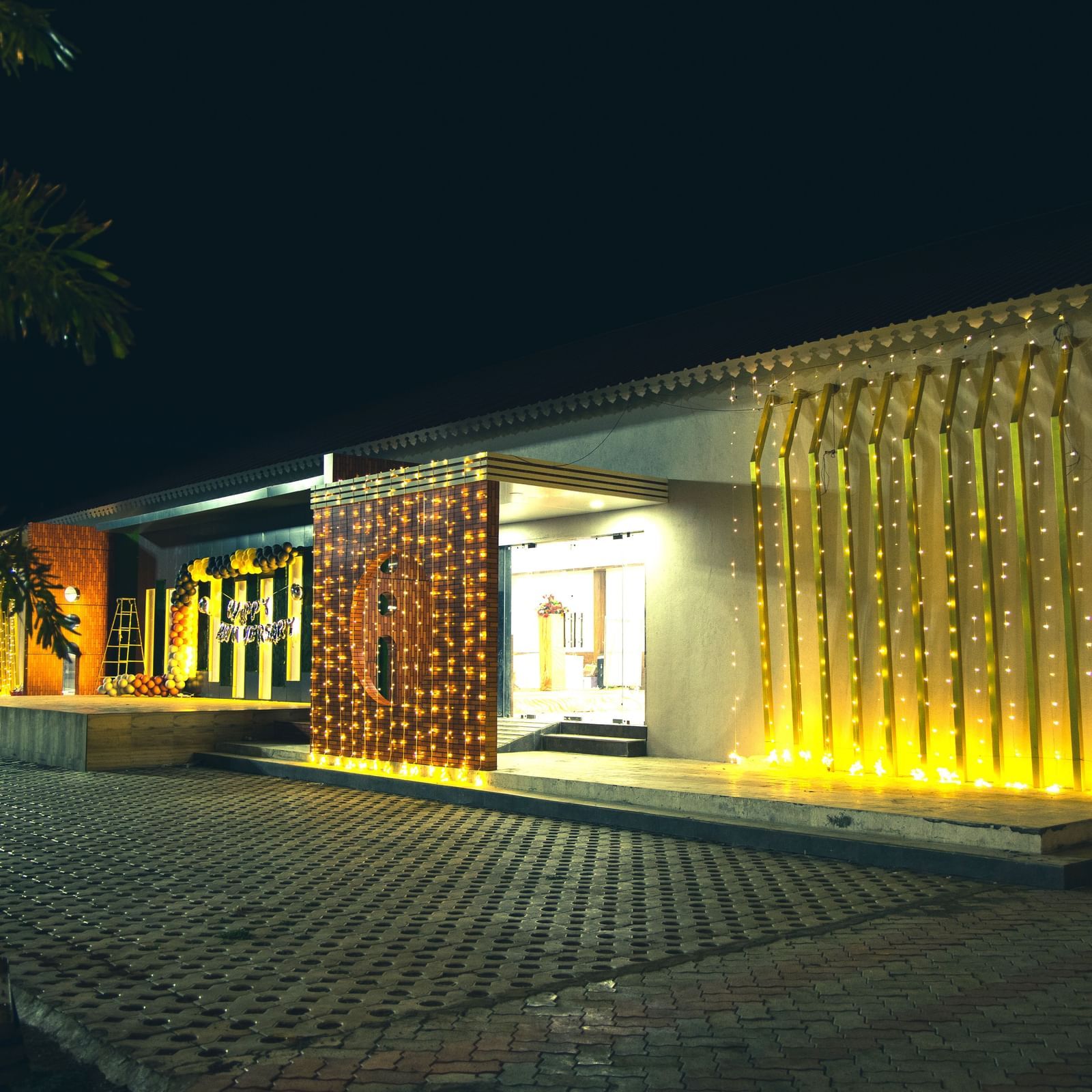 A night view of the entry of an event space in Daksh Eden Greenz decorated with fairy lights and a 'happy birthday' balloon arrangement on the wall.