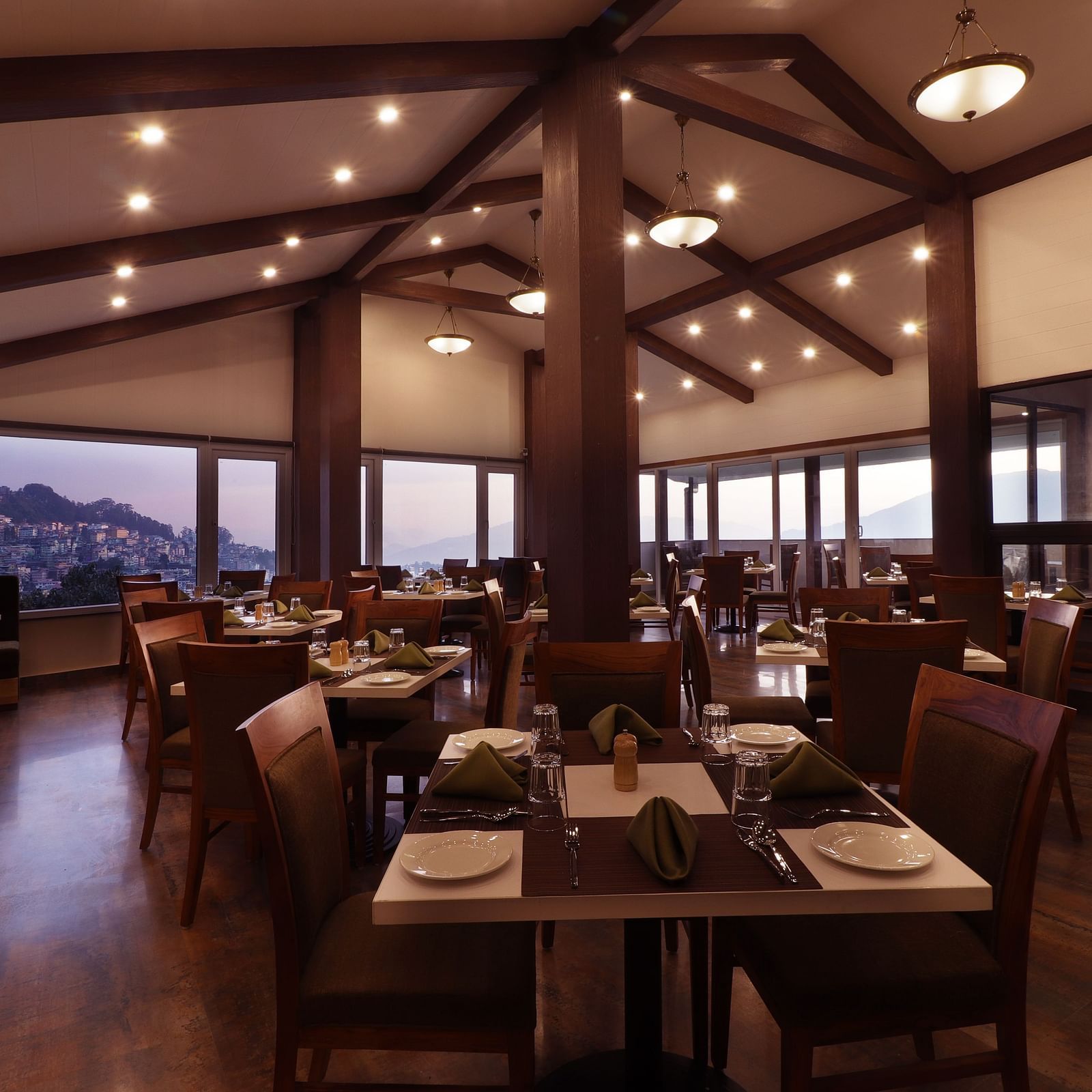 A modern styled rooftop dining space featuring wooden furniture and arranged table set with tableware and folded napkins and huge windows that opens to hilly cityscape at the backdrop.