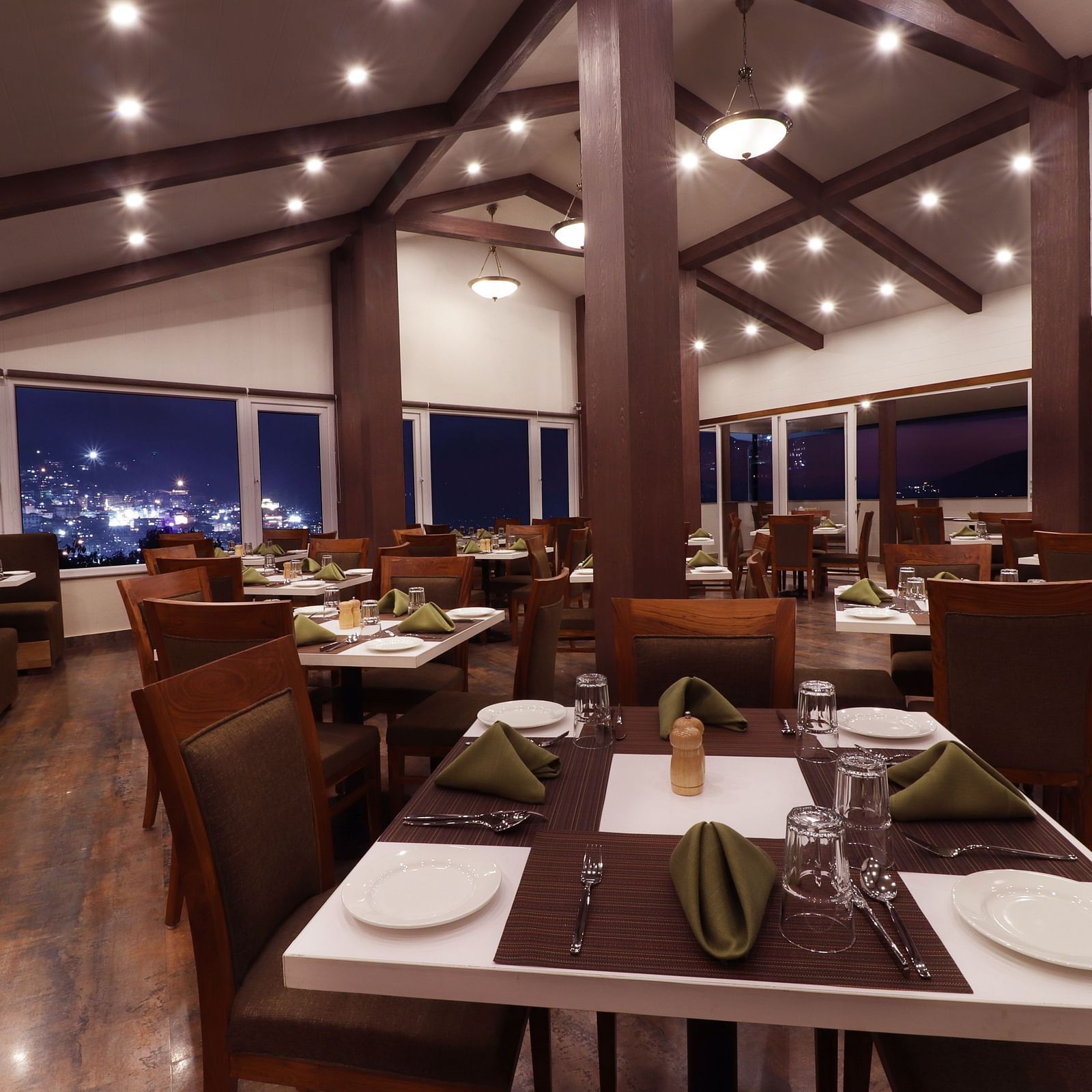 A close up view of the rooftop dining hall at Denzong Shangrila Lords Inn Gangtok featuring wooden furnishing, arranged table set with tableware and large windows opening to the evening view of illuminated town.