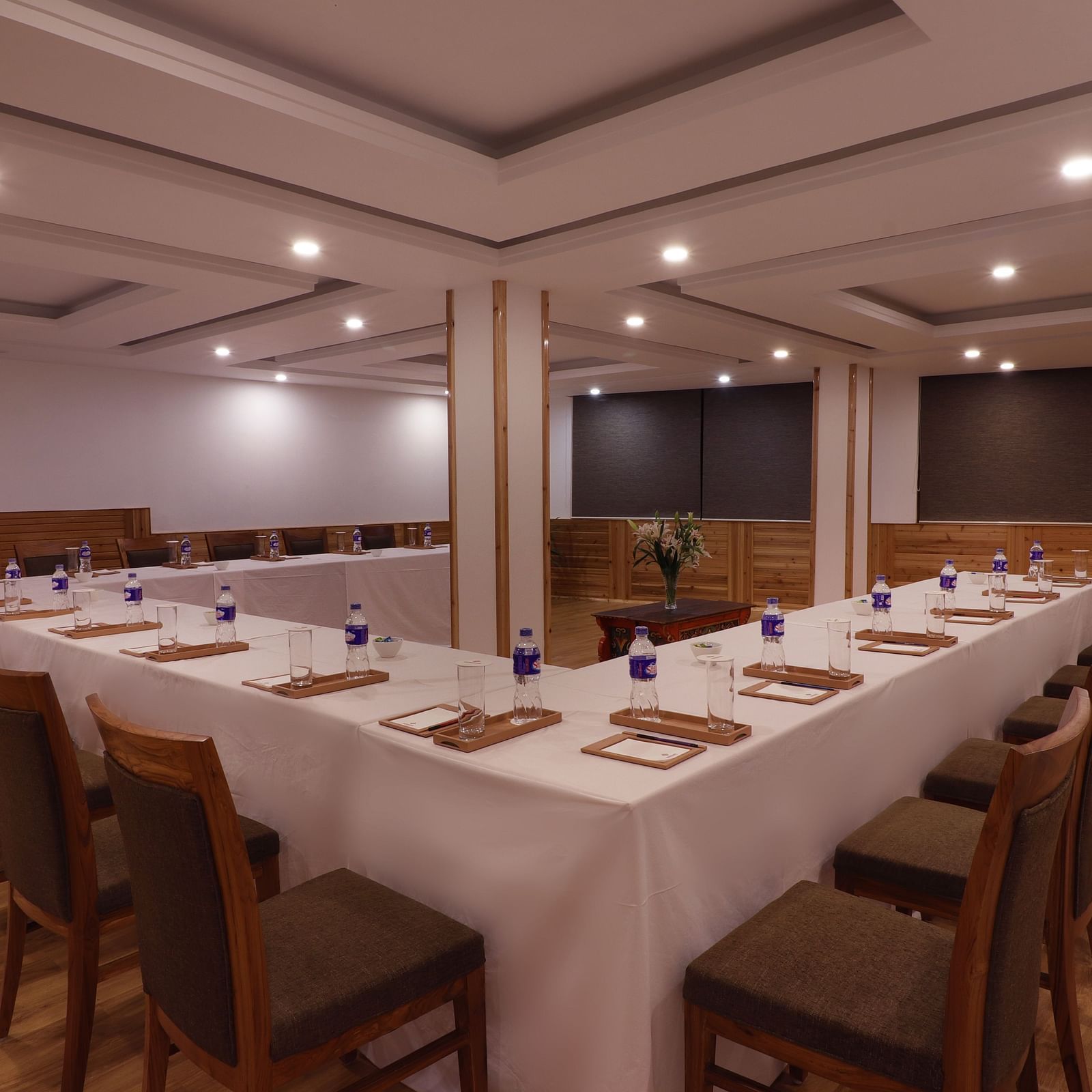 A symmetrical view of arranged elongated table and wooden chairs under a dimly lit spacious banquet hall at Denzong Shangrila Lords Inn Gangtok.
