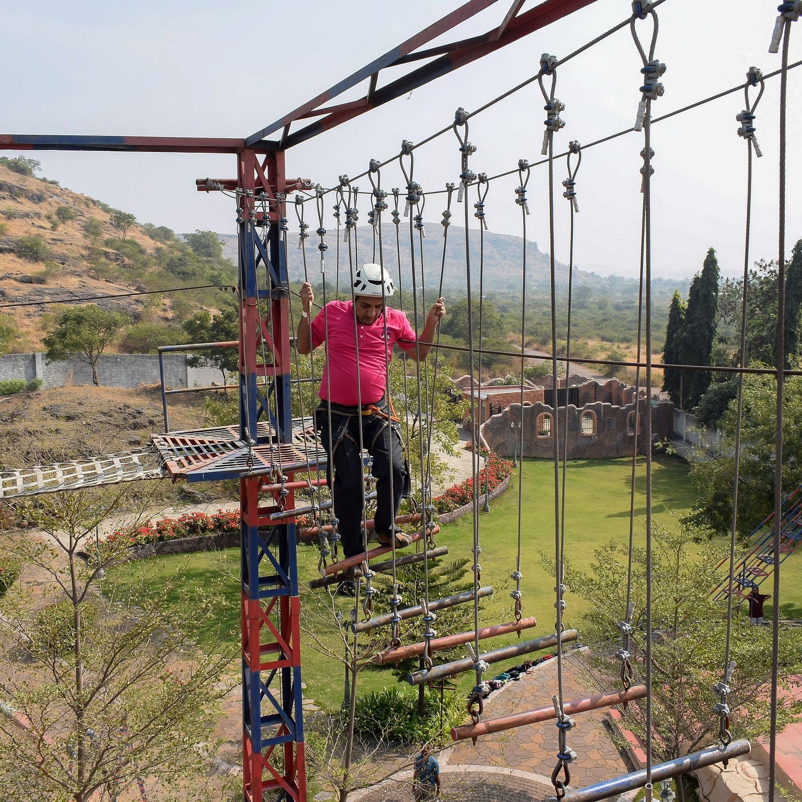A person traversing a high rope obstacle course at an outdoor adventure park, scenic hills in the background | Diamond Parks, Pune