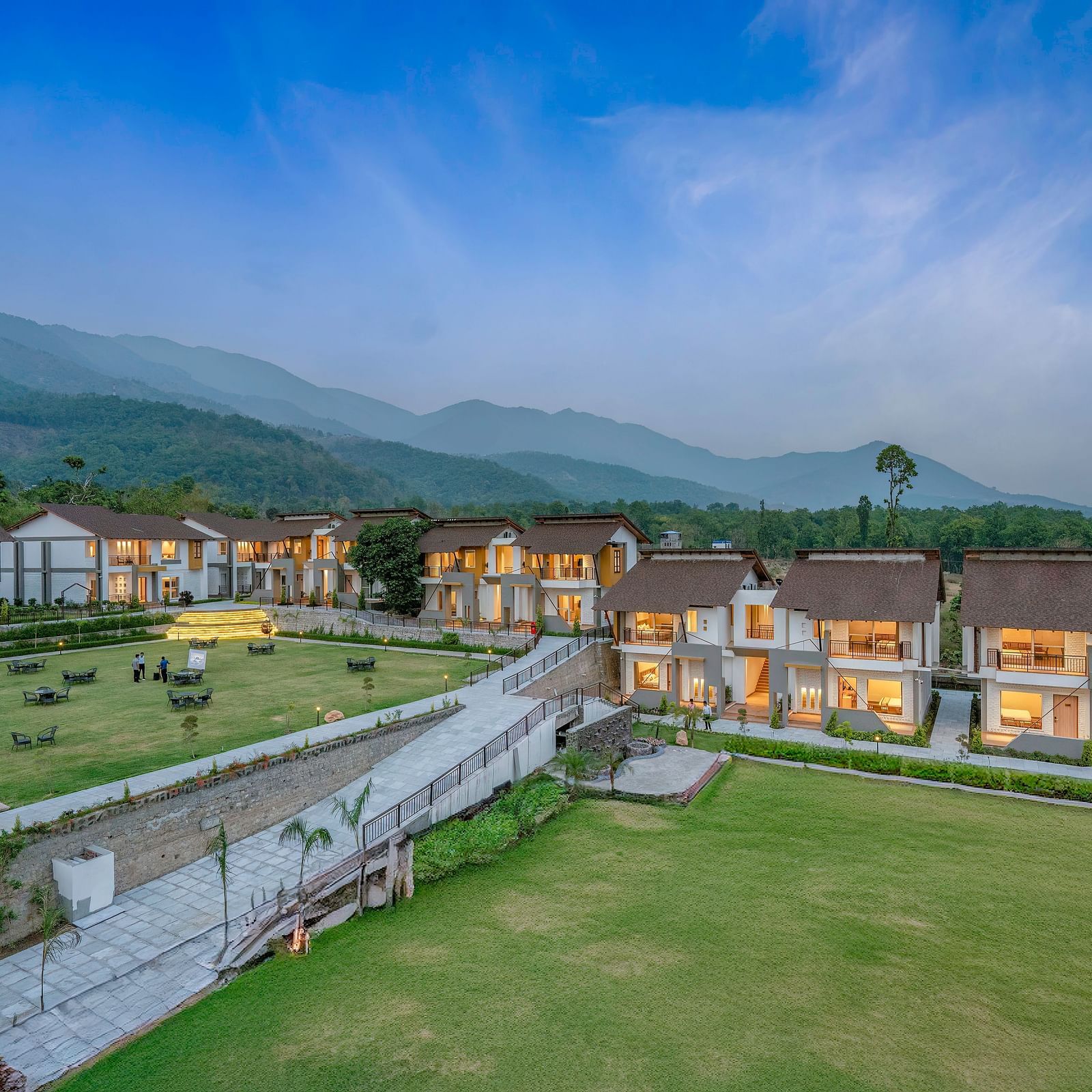 Shot of a resort with multiple buildings, a swimming pool, and a large lawn, surrounded by mountains.