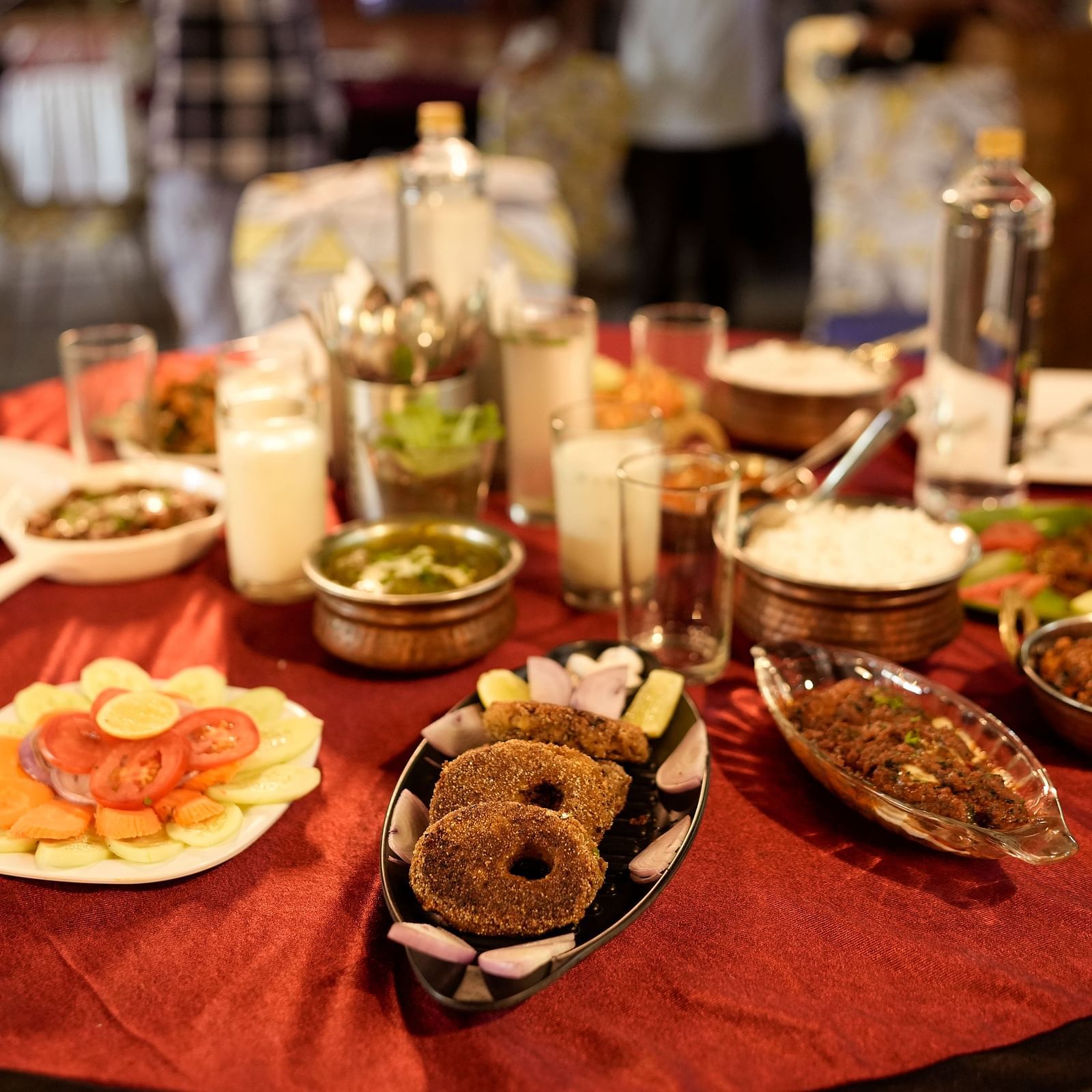 A round table covered with a red tablecloth, displaying various dishes of South Asian cuisine, including curry, lentils, and salads. There are also multiple glasses and a bottle on the table. Plates are set for diners.